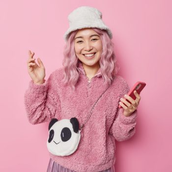 Positive young pretty Asian woman with pink hair wears hat and jacket carries panda bag holds smartphone smiles positively glad to receive message from boyfriend isolated over pink background